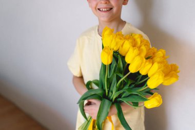 Bir çocuk elinde büyük bir buket sarı lale ile evde duruyor, yüzünde kocaman bir gülümsemeyle mutluluk saçıyor. Parlak atmosfer neşe ve sıcaklığı yansıtır. Anneler Günü ve Bahar Konsepti