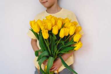 Bir çocuk elinde büyük bir buket sarı lale ile evde duruyor, yüzünde kocaman bir gülümsemeyle mutluluk saçıyor. Parlak atmosfer neşe ve sıcaklığı yansıtır. Anneler Günü ve Bahar Konsepti