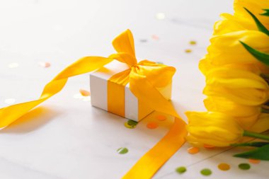 Uzun sarı kurdeleli hediye kutusu ve açık arkaplanda laleler.