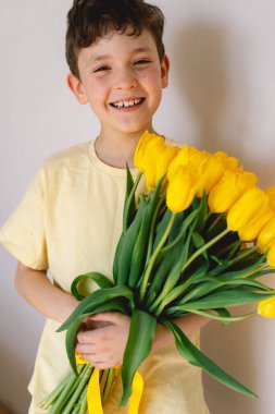 Bir çocuk elinde büyük bir buket sarı lale ile evde duruyor, yüzünde kocaman bir gülümsemeyle mutluluk saçıyor. Parlak atmosfer neşe ve sıcaklığı yansıtır. Anneler Günü ve Bahar Konsepti