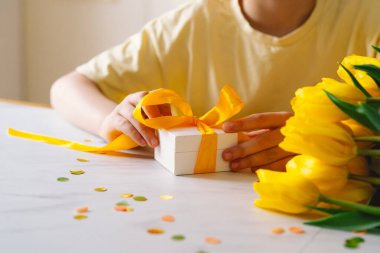 Bir çocuk mutfak masasında oturmuş, anneler günü hediyesinde fiyonk bağlıyor. Güzel sarı laleler onu sarıp, neşeli bir atmosfer yaratıyor..