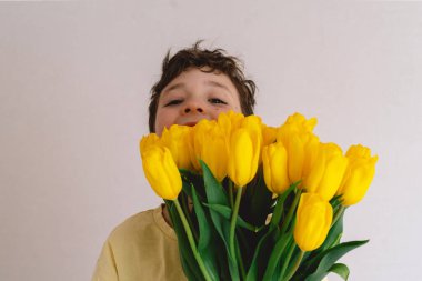 Bir çocuk elinde büyük bir buket sarı lale ile evde duruyor, yüzünde kocaman bir gülümsemeyle mutluluk saçıyor. Parlak atmosfer neşe ve sıcaklığı yansıtır. Anneler Günü ve Bahar Konsepti