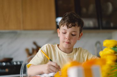 Bir çocuk mutfak masasında oturmuş, özenle anneler günü kartı hazırlıyor ve 
