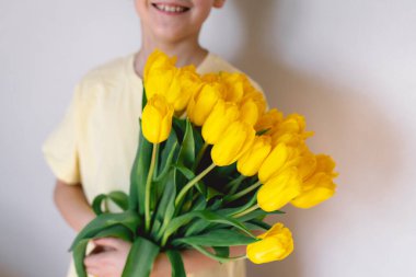 Bir çocuk elinde büyük bir buket sarı lale ile evde duruyor, yüzünde kocaman bir gülümsemeyle mutluluk saçıyor. Parlak atmosfer neşe ve sıcaklığı yansıtır. Anneler Günü ve Bahar Konsepti