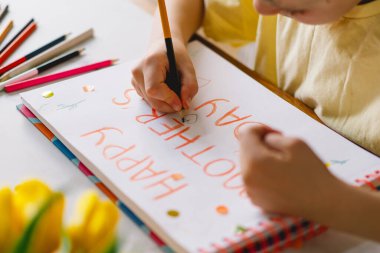 Bir çocuk mutfak masasında oturmuş, özenle anneler günü kartı hazırlıyor ve 