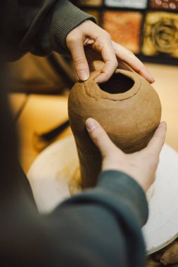 Çömlek atölyesinde kilden bir tencereyi ustalıkla şekillendiren eller. Atölye yaratıcılığı teşvik ediyor ve yeni başlayanlara rahat bir ortamda çömlek teknikleri öğretmeye odaklanıyor..