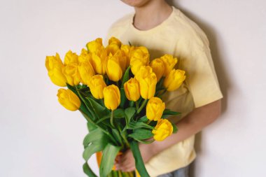 Bir çocuk elinde büyük bir buket sarı lale ile evde duruyor, yüzünde kocaman bir gülümsemeyle mutluluk saçıyor. Parlak atmosfer neşe ve sıcaklığı yansıtır. Anneler Günü ve Bahar Konsepti