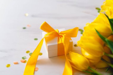 Uzun sarı kurdeleli hediye kutusu ve açık arkaplanda laleler.