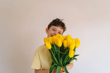 Bir çocuk elinde büyük bir buket sarı lale ile evde duruyor, yüzünde kocaman bir gülümsemeyle mutluluk saçıyor. Parlak atmosfer neşe ve sıcaklığı yansıtır. Anneler Günü ve Bahar Konsepti