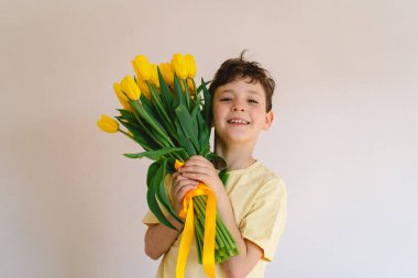 Bir çocuk elinde büyük bir buket sarı lale ile evde duruyor, yüzünde kocaman bir gülümsemeyle mutluluk saçıyor. Parlak atmosfer neşe ve sıcaklığı yansıtır. Anneler Günü ve Bahar Konsepti