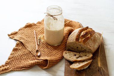 Çıtır ekşi ekmek, sıcak bir mutfak atmosferinde kırsal bir bez ve ahşap bir tahtanın eşlik ettiği etkin marş başlatıcısıyla dolu cam bir kavanozun yanında oturur..