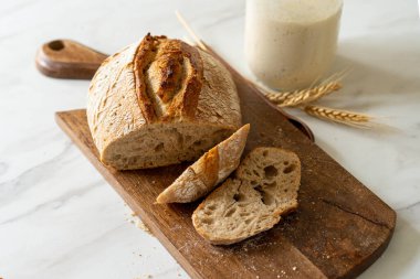Çıtır ekşi ekmek, sıcak bir mutfak atmosferinde kırsal bir bez ve ahşap bir tahtanın eşlik ettiği etkin marş başlatıcısıyla dolu cam bir kavanozun yanında oturur..