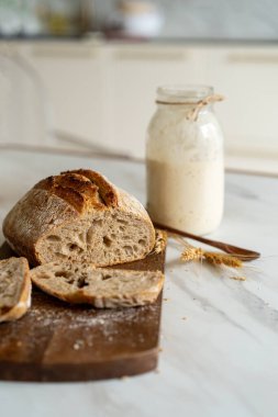 Çıtır ekşi ekmek, sıcak bir mutfak atmosferinde kırsal bir bez ve ahşap bir tahtanın eşlik ettiği etkin marş başlatıcısıyla dolu cam bir kavanozun yanında oturur..