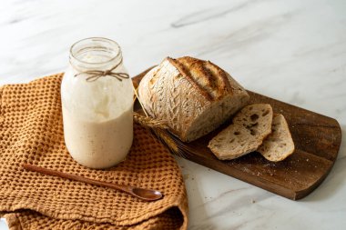 Çıtır ekşi ekmek, sıcak bir mutfak atmosferinde kırsal bir bez ve ahşap bir tahtanın eşlik ettiği etkin marş başlatıcısıyla dolu cam bir kavanozun yanında oturur..