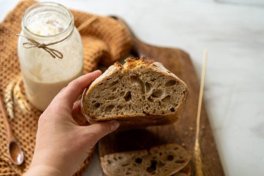 Çıtır ekşi ekmek, sıcak bir mutfak atmosferinde kırsal bir bez ve ahşap bir tahtanın eşlik ettiği etkin marş başlatıcısıyla dolu cam bir kavanozun yanında oturur..