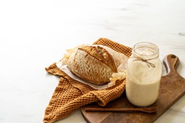 Çıtır ekşi ekmek, sıcak bir mutfak atmosferinde kırsal bir bez ve ahşap bir tahtanın eşlik ettiği etkin marş başlatıcısıyla dolu cam bir kavanozun yanında oturur..