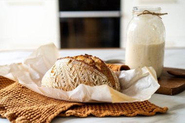 Çıtır ekşi ekmek, sıcak bir mutfak atmosferinde kırsal bir bez ve ahşap bir tahtanın eşlik ettiği etkin marş başlatıcısıyla dolu cam bir kavanozun yanında oturur..