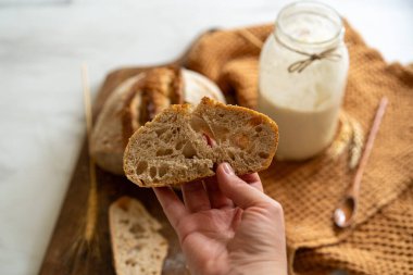Çıtır ekşi ekmek, sıcak bir mutfak atmosferinde kırsal bir bez ve ahşap bir tahtanın eşlik ettiği etkin marş başlatıcısıyla dolu cam bir kavanozun yanında oturur..