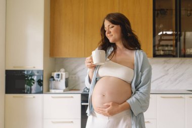 Sıcak bir mutfakta hamile bir kadın hamile karnını nazikçe tutuyor. Huzurlu bir ortamda yaklaşan anneliği yansıtıyor.