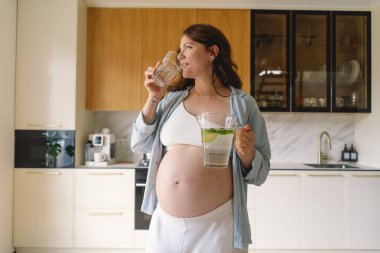 Günümüz mutfağında, neşeli bir suratı olan hamile bir kadın bir sürahi dolusu su ve bardak tutuyor. Parlak ışık, o su içmekten zevk alırken onun ışıl ışıl gülümsemesini arttırır..
