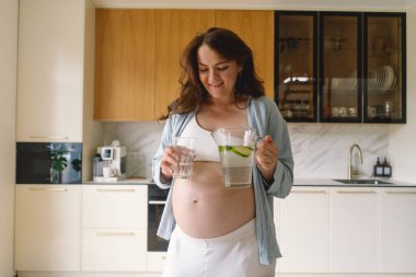 Günümüz mutfağında, neşeli bir suratı olan hamile bir kadın bir sürahi dolusu su ve bardak tutuyor. Parlak ışık, o su içmekten zevk alırken onun ışıl ışıl gülümsemesini arttırır..