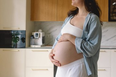 Sıcak bir mutfakta hamile bir kadın hamile karnını nazikçe tutuyor. Huzurlu bir ortamda yaklaşan anneliği yansıtıyor.