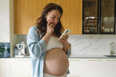 Hamile kadın çağdaş bir mutfakta duruyor, akıllı telefonuna odaklanmış. Doğal ışık mekanı aydınlatır, rahat ve şık bir ortam sunar..