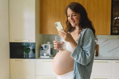 Hamile kadın çağdaş bir mutfakta duruyor, akıllı telefonuna odaklanmış. Doğal ışık mekanı aydınlatır, rahat ve şık bir ortam sunar..