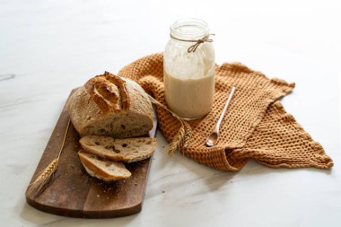 Çıtır ekşi ekmek, sıcak bir mutfak atmosferinde kırsal bir bez ve ahşap bir tahtanın eşlik ettiği etkin marş başlatıcısıyla dolu cam bir kavanozun yanında oturur..