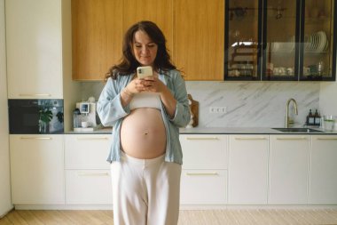 Hamile kadın çağdaş bir mutfakta duruyor, akıllı telefonuna odaklanmış. Doğal ışık mekanı aydınlatır, rahat ve şık bir ortam sunar..