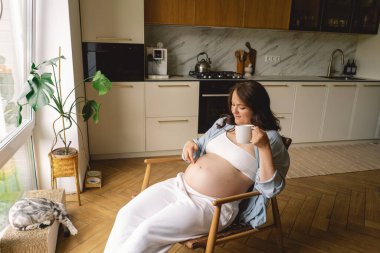 Sıcak bir mutfakta hamile bir kadın hamile karnını nazikçe tutuyor. Huzurlu bir ortamda yaklaşan anneliği yansıtıyor.