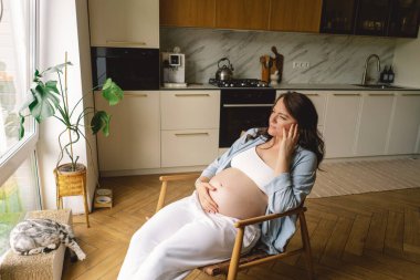 Sıcak bir mutfakta hamile bir kadın hamile karnını nazikçe tutuyor. Huzurlu bir ortamda yaklaşan anneliği yansıtıyor.