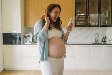 Hamile bir kadın şık bir mutfakta elinde fincanla telefonda konuşurken gülümsüyor. Alan ahşap dolaplar ve zarif aletler içeriyor, rahat bir atmosfer yaratıyor..