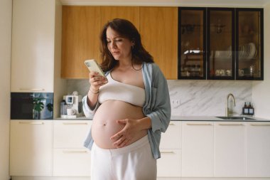 Üç aylık hamile bir kadın, acı dolu bir ifadeyle karnını tutuyor. Gündüz saatlerinde modern bir mutfak ortamında duran hamile bir kadın rahatsız oluyor.