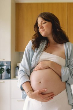 Sıcak bir mutfakta hamile bir kadın hamile karnını nazikçe tutuyor. Huzurlu bir ortamda yaklaşan anneliği yansıtıyor.