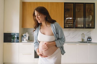 Üç aylık hamile bir kadın, acı dolu bir ifadeyle karnını tutuyor. Gündüz saatlerinde modern bir mutfak ortamında duran hamile bir kadın rahatsız oluyor.