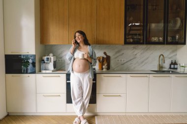 Hamile bir kadın şık bir mutfakta elinde fincanla telefonda konuşurken gülümsüyor. Alan ahşap dolaplar ve zarif aletler içeriyor, rahat bir atmosfer yaratıyor..