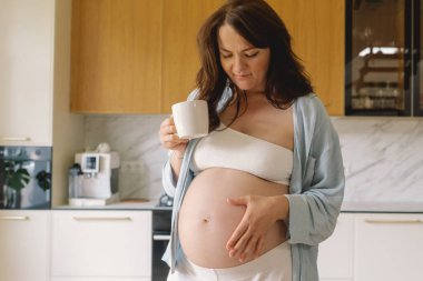 Sıcak bir mutfakta hamile bir kadın hamile karnını nazikçe tutuyor. Huzurlu bir ortamda yaklaşan anneliği yansıtıyor.