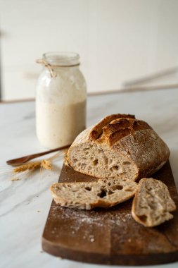 Çıtır ekşi ekmek, sıcak bir mutfak atmosferinde kırsal bir bez ve ahşap bir tahtanın eşlik ettiği etkin marş başlatıcısıyla dolu cam bir kavanozun yanında oturur..
