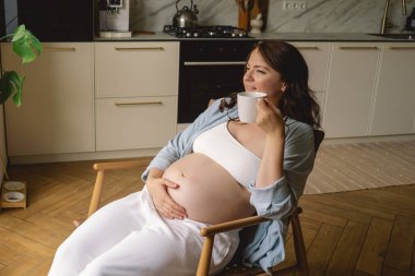 Sıcak bir mutfakta hamile bir kadın hamile karnını nazikçe tutuyor. Huzurlu bir ortamda yaklaşan anneliği yansıtıyor.