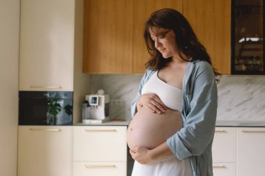 Sıcak bir mutfakta hamile bir kadın hamile karnını nazikçe tutuyor. Huzurlu bir ortamda yaklaşan anneliği yansıtıyor.