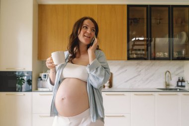 Hamile bir kadın şık bir mutfakta elinde fincanla telefonda konuşurken gülümsüyor. Alan ahşap dolaplar ve zarif aletler içeriyor, rahat bir atmosfer yaratıyor..