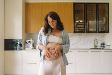 Sıcak bir mutfakta hamile bir kadın hamile karnını nazikçe tutuyor. Huzurlu bir ortamda yaklaşan anneliği yansıtıyor.