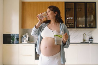 Günümüz mutfağında, neşeli bir suratı olan hamile bir kadın bir sürahi dolusu su ve bardak tutuyor. Parlak ışık, o su içmekten zevk alırken onun ışıl ışıl gülümsemesini arttırır..
