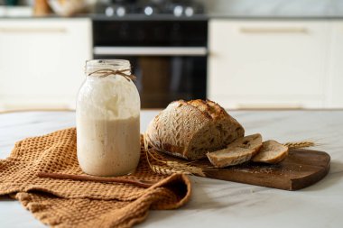 Çıtır ekşi ekmek, sıcak bir mutfak atmosferinde kırsal bir bez ve ahşap bir tahtanın eşlik ettiği etkin marş başlatıcısıyla dolu cam bir kavanozun yanında oturur..