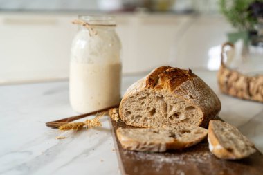 Çıtır ekşi ekmek, sıcak bir mutfak atmosferinde kırsal bir bez ve ahşap bir tahtanın eşlik ettiği etkin marş başlatıcısıyla dolu cam bir kavanozun yanında oturur..