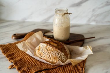 Çıtır ekşi ekmek, sıcak bir mutfak atmosferinde kırsal bir bez ve ahşap bir tahtanın eşlik ettiği etkin marş başlatıcısıyla dolu cam bir kavanozun yanında oturur..