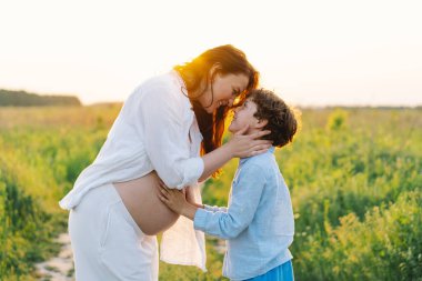 Hamile bir kadın yemyeşil bir tarlada duruyor, nazikçe bebeğini tutuyor. Oğlu ona yaslanıyor, uzun otlar ve sıcak bir gün batımı arasında hassas bir anı paylaşıyor..