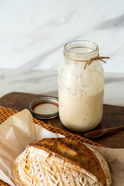 Çıtır ekşi ekmek, sıcak bir mutfak atmosferinde kırsal bir bez ve ahşap bir tahtanın eşlik ettiği etkin marş başlatıcısıyla dolu cam bir kavanozun yanında oturur..