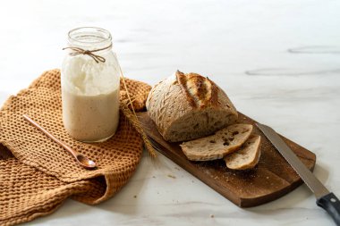 Çıtır ekşi ekmek, sıcak bir mutfak atmosferinde kırsal bir bez ve ahşap bir tahtanın eşlik ettiği etkin marş başlatıcısıyla dolu cam bir kavanozun yanında oturur..